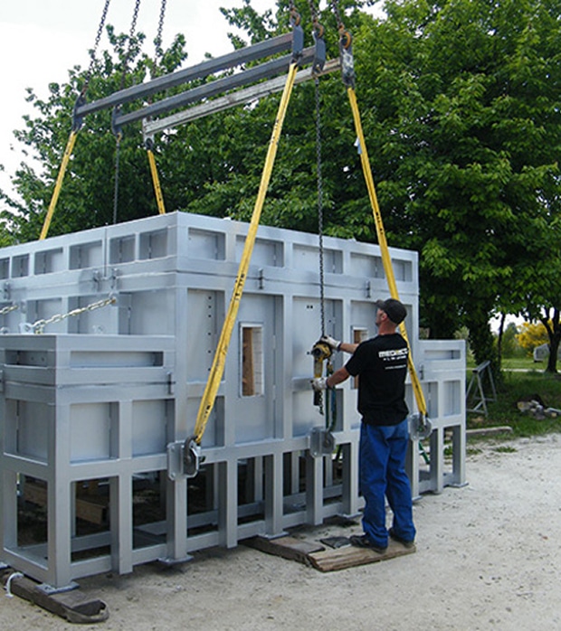 Levage sécurisé d'une structure industrielle lourde Structure industrielle grise soulevée par une traverse et quatre élingues jaunes. Un opérateur REDECKT guide la lourde charge lors du levage.